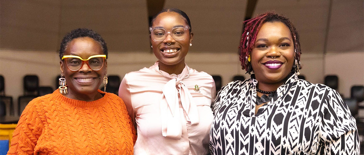 Three African American women smile. The youngest of the three, in the center, wears a pin. 