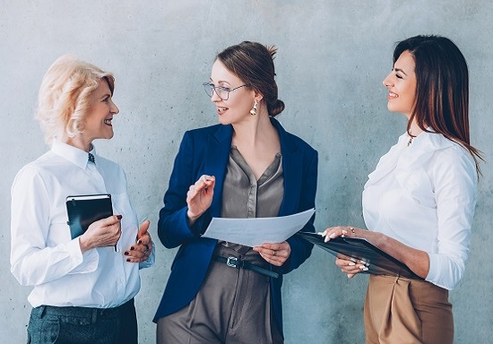 Tres mujeres de negocios vestidas elegantemente hablando de un proyecto y con carpetas en la mano.