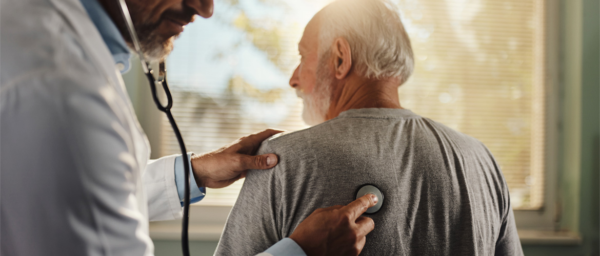 doctor in white coat holds stethescope on the back of an elderly patient in sunny room