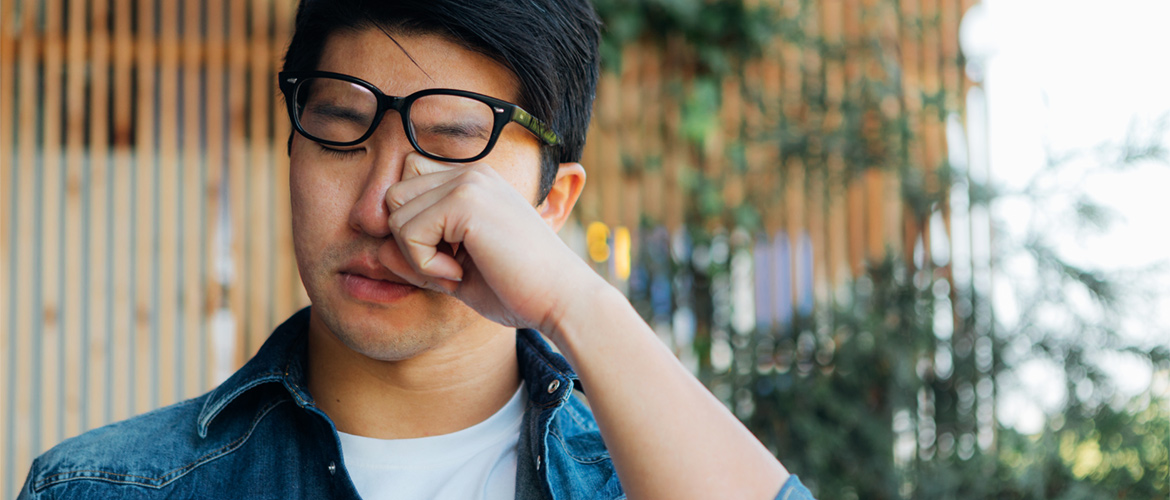 Man outside in denim shirt pushes up black glasses to rub left eye with hand