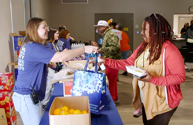 Woman standing behind a table hands a blue bag to another woman on the other side of the table