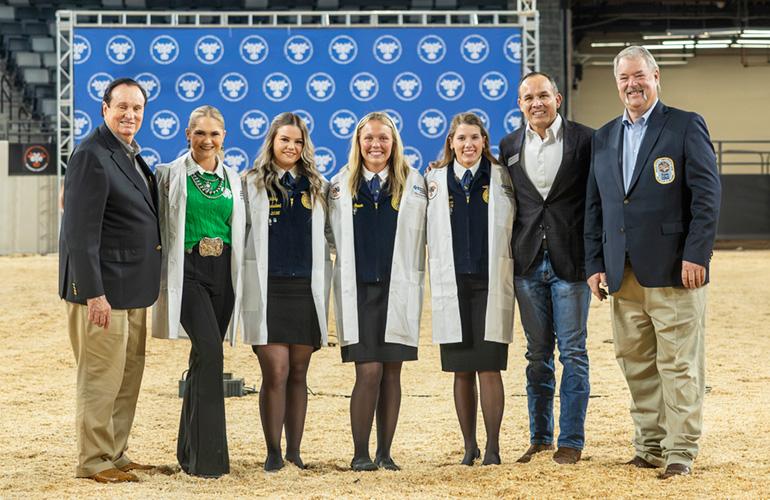 Four young women and three men in suits stand for posed photo in arena with dirt ground
