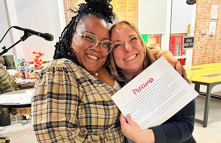 Two women smile in a classroom with the woman on the right holding a certificate