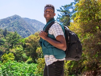 Joven afroamericano con ropa de senderismo y mochila con una montaña y árboles de fondo