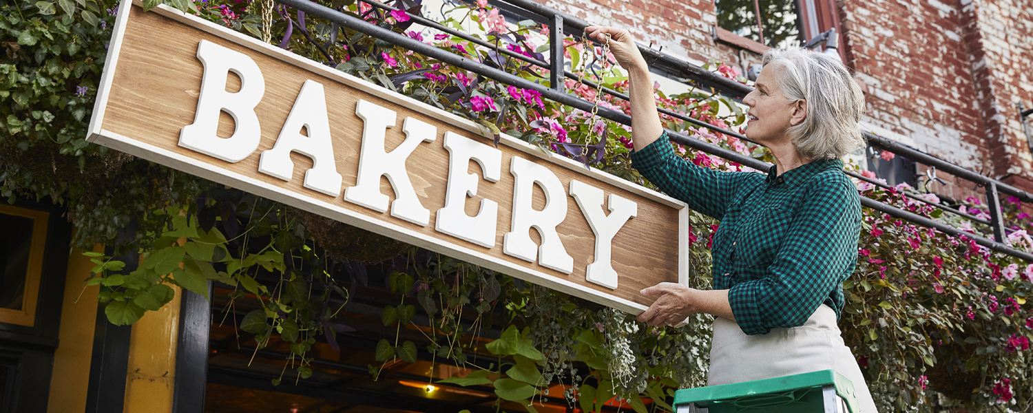 A woman standing on a ladder hangs a sign that says "BAKERY" outside