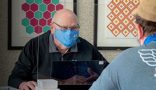 Two men in masks separated by plexiglass discuss health care.