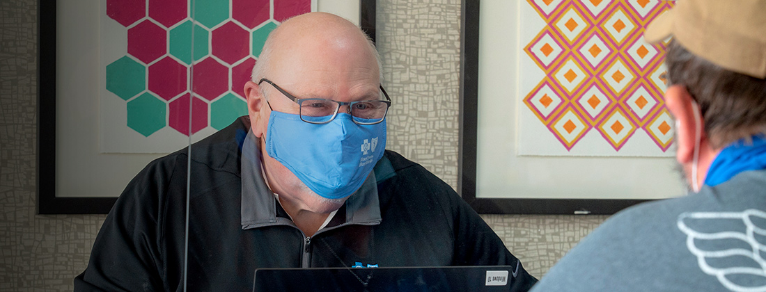 Dos hombres con mascarillas separados por plexiglás dialogan sobre la atención médica.