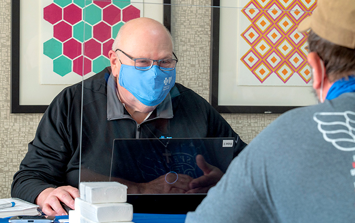 Two men in masks separated by plexiglass discuss health care 