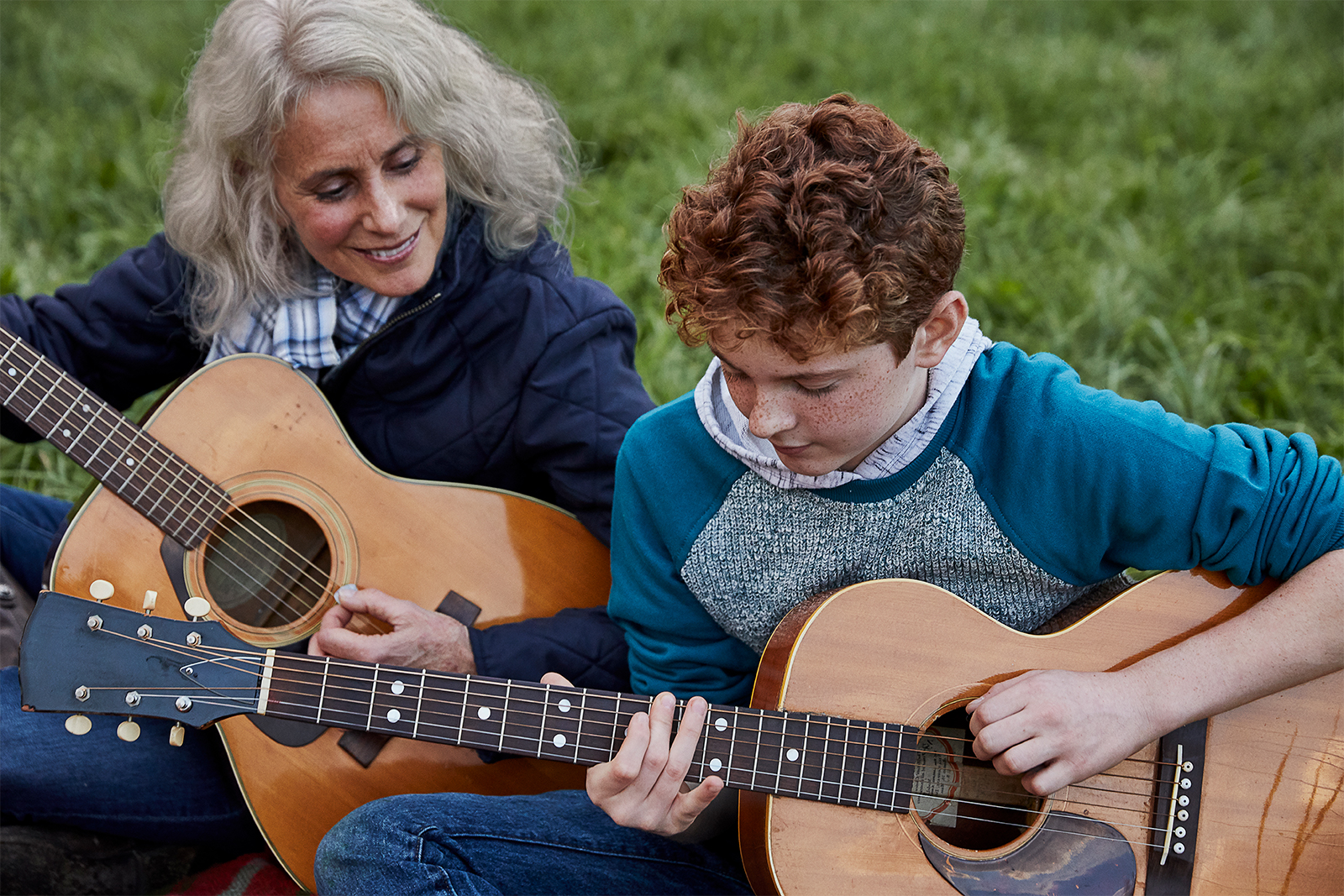 senior woman sitting in the grass teaching her grandson to play the guitar.  